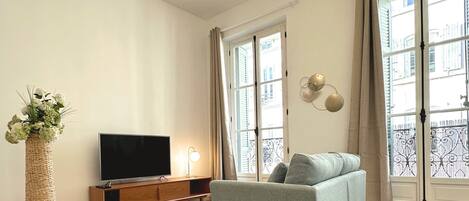 A spacious living room with a modern sofa, a flat-screen TV, a wooden TV stand, a decorative vase, and large windows with a view of buildings.