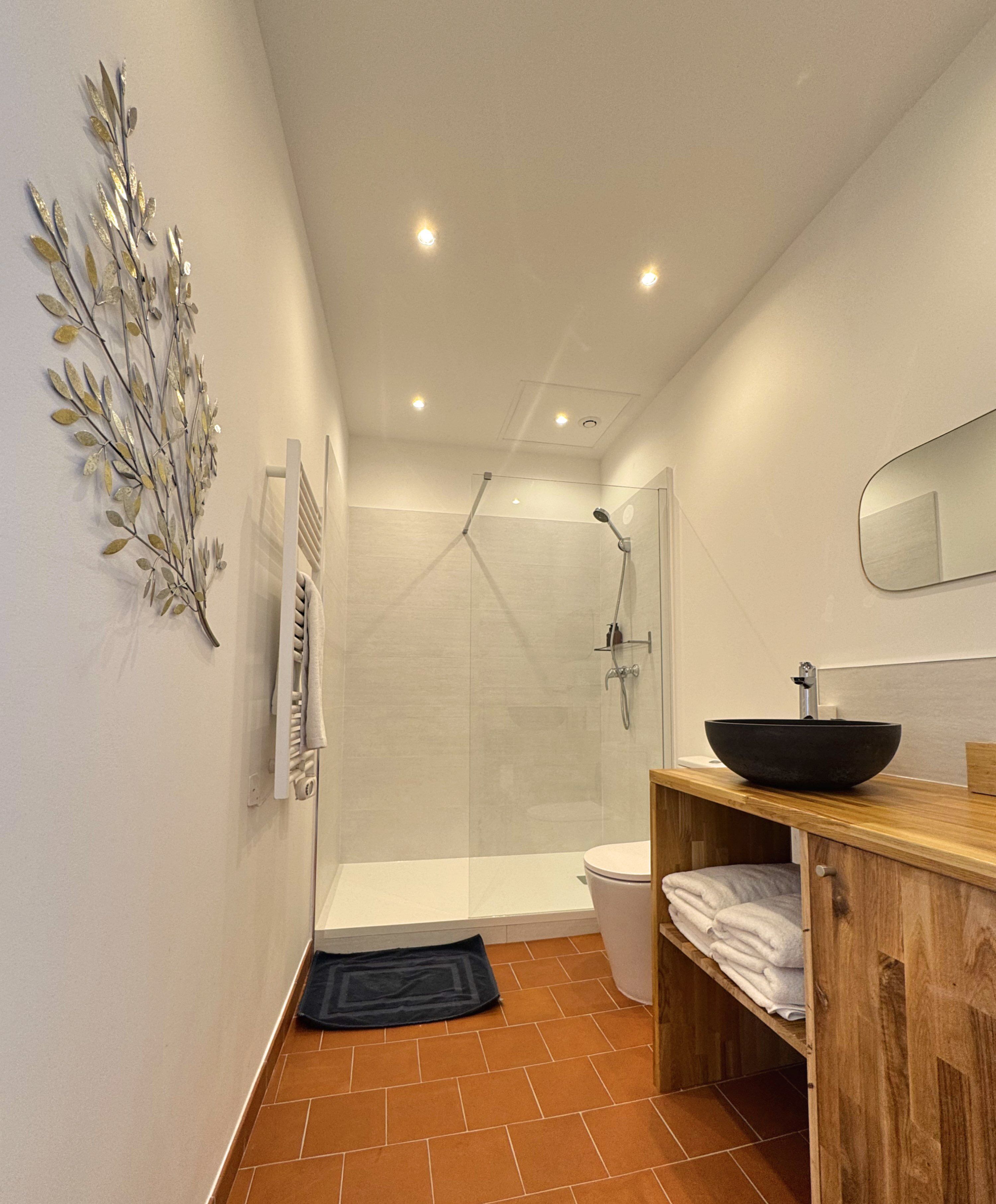 A modern bathroom with a glass-enclosed shower, a wooden vanity with a black basin, and a decorative metal wall piece.