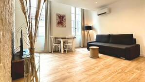 A spacious living area with a wooden floor, a black sofa, a dining table with chairs, and a vase with tall grasses. - Les Appartements du Vieux-Port (Marseille)