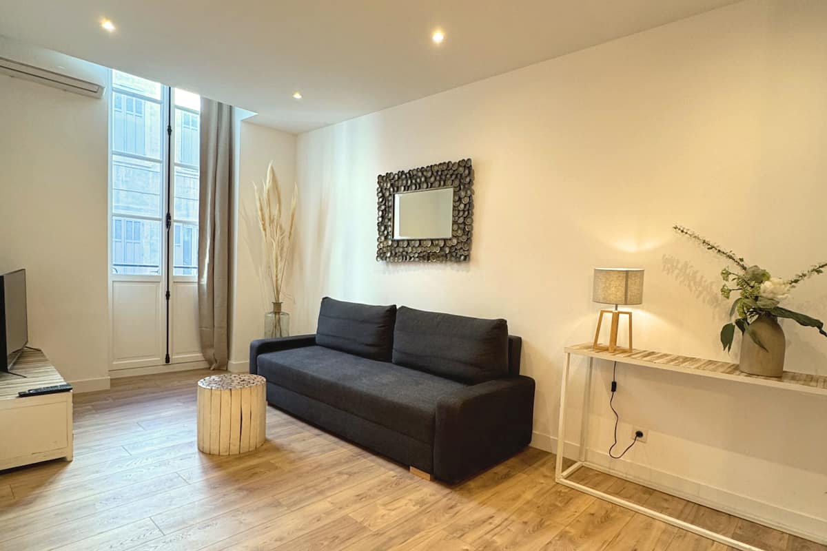 A modern living room with a dark grey sofa, a small round table, a white cabinet, and a large window with curtains.