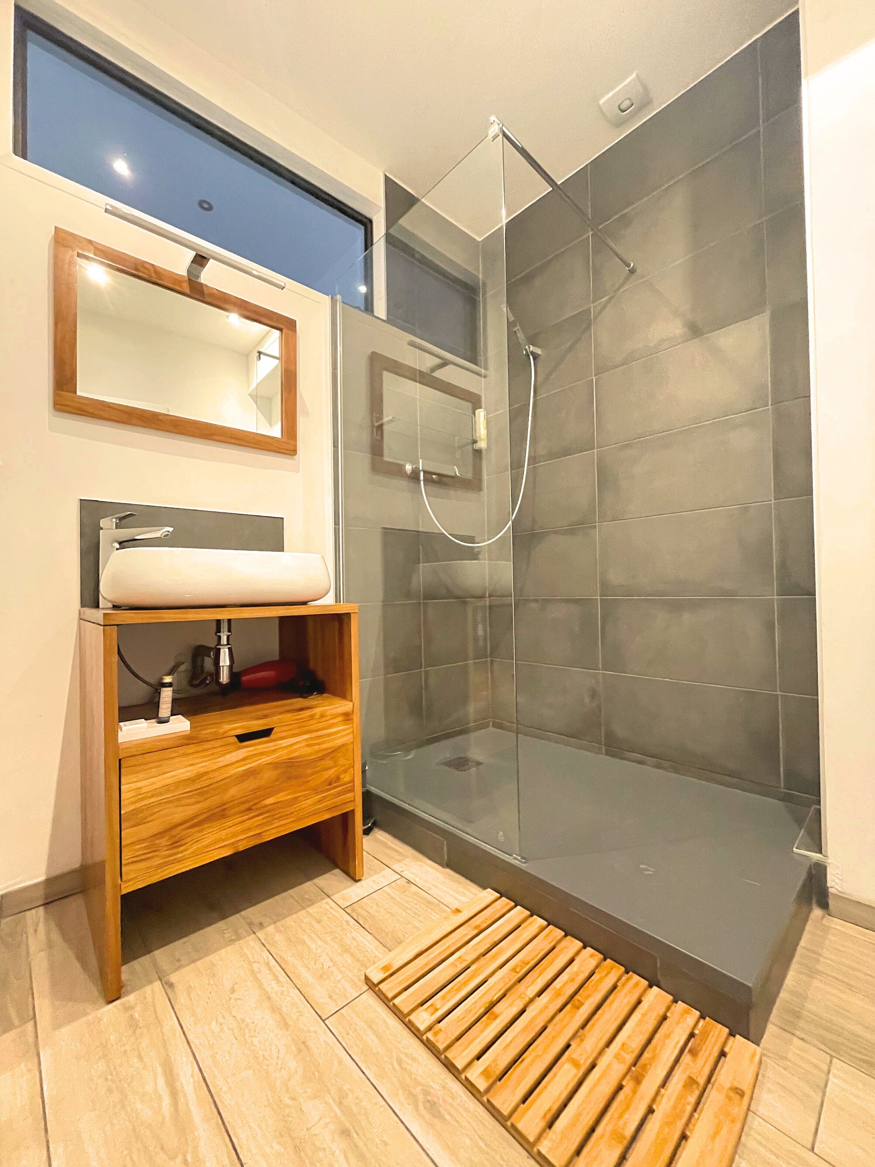 A modern bathroom with a glass-enclosed shower, a wooden vanity with a mirror, and a tiled floor.