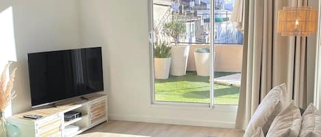 A modern living room with a large flat-screen TV, a sofa, a wicker side table, and a view of a cityscape through large windows.