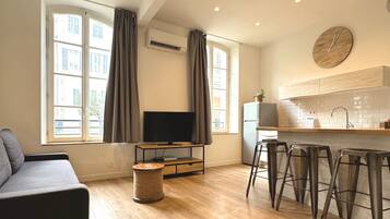 A modern living room with a sofa, a TV, a kitchen area with bar stools, and a window with curtains.