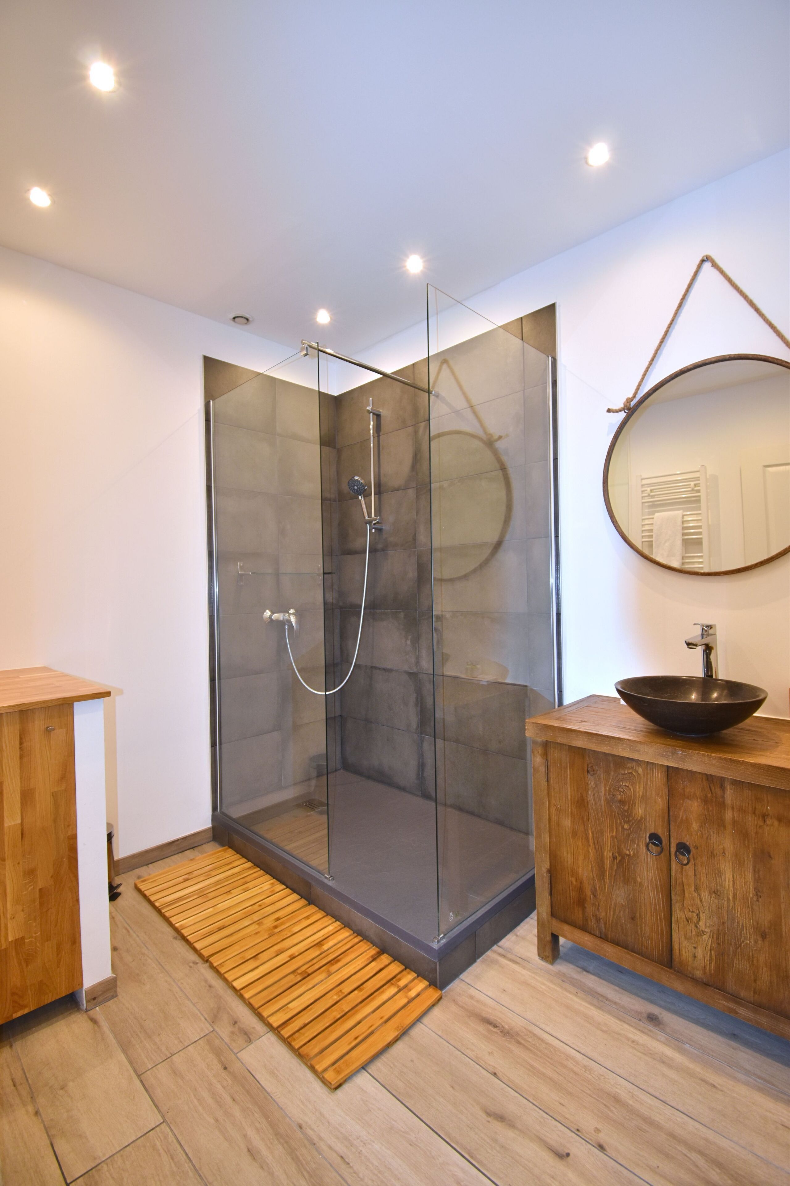 A modern bathroom with a glass-enclosed shower, wooden vanity, and a round mirror.