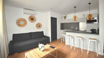 A modern living room with a dark grey sofa, a wooden coffee table with a vase of flowers, and a kitchen area with a white countertop, bar stools, and pendant lighting.