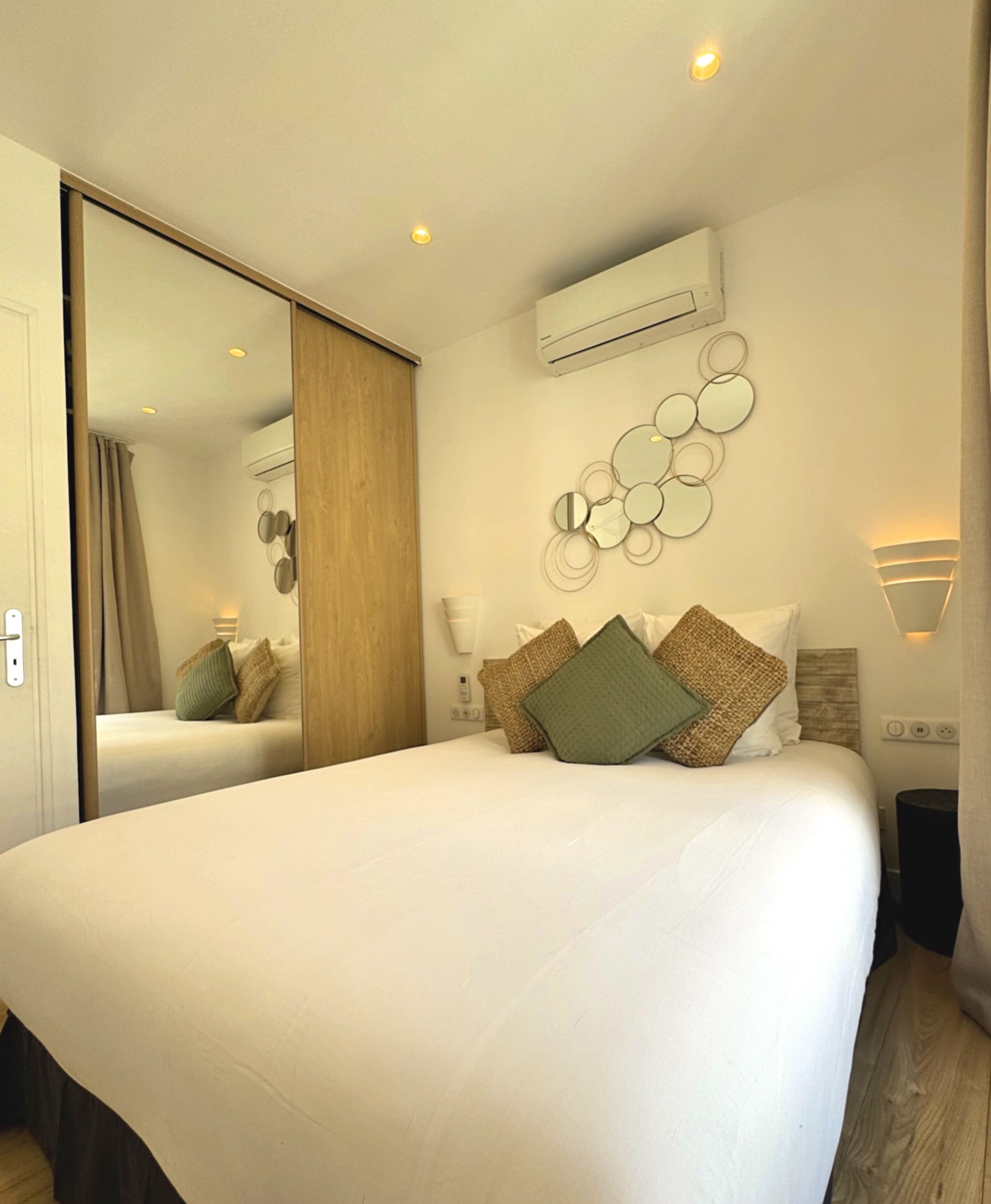 A modern hotel room with a large bed, a mirror, an air conditioning unit, and a wall-mounted light fixture.