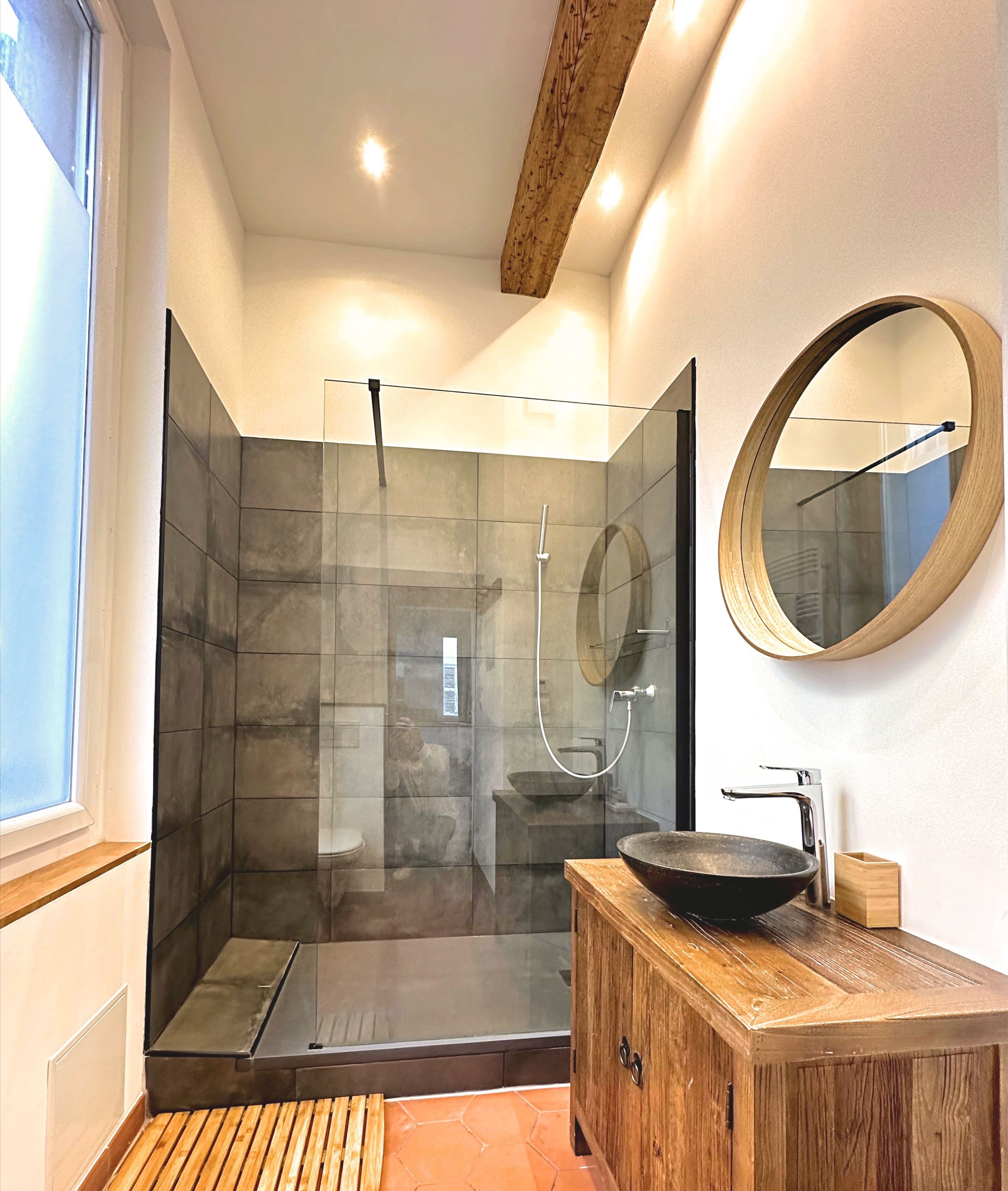 Un baño moderno con ducha de vidrio, tocador de madera con lavabo y espejo redondo.