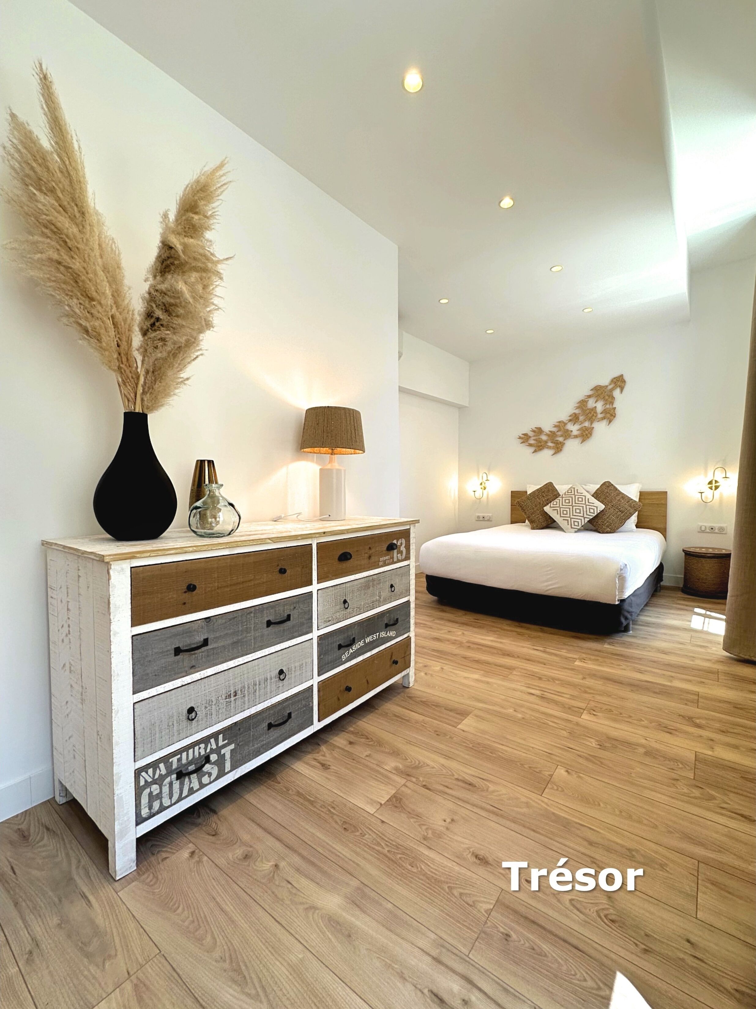 Un dormitorio moderno con piso de madera, una cama blanca, un mesita de noche, un aparador con varios cajones, una lámpara y hierba de pampa en un florero.