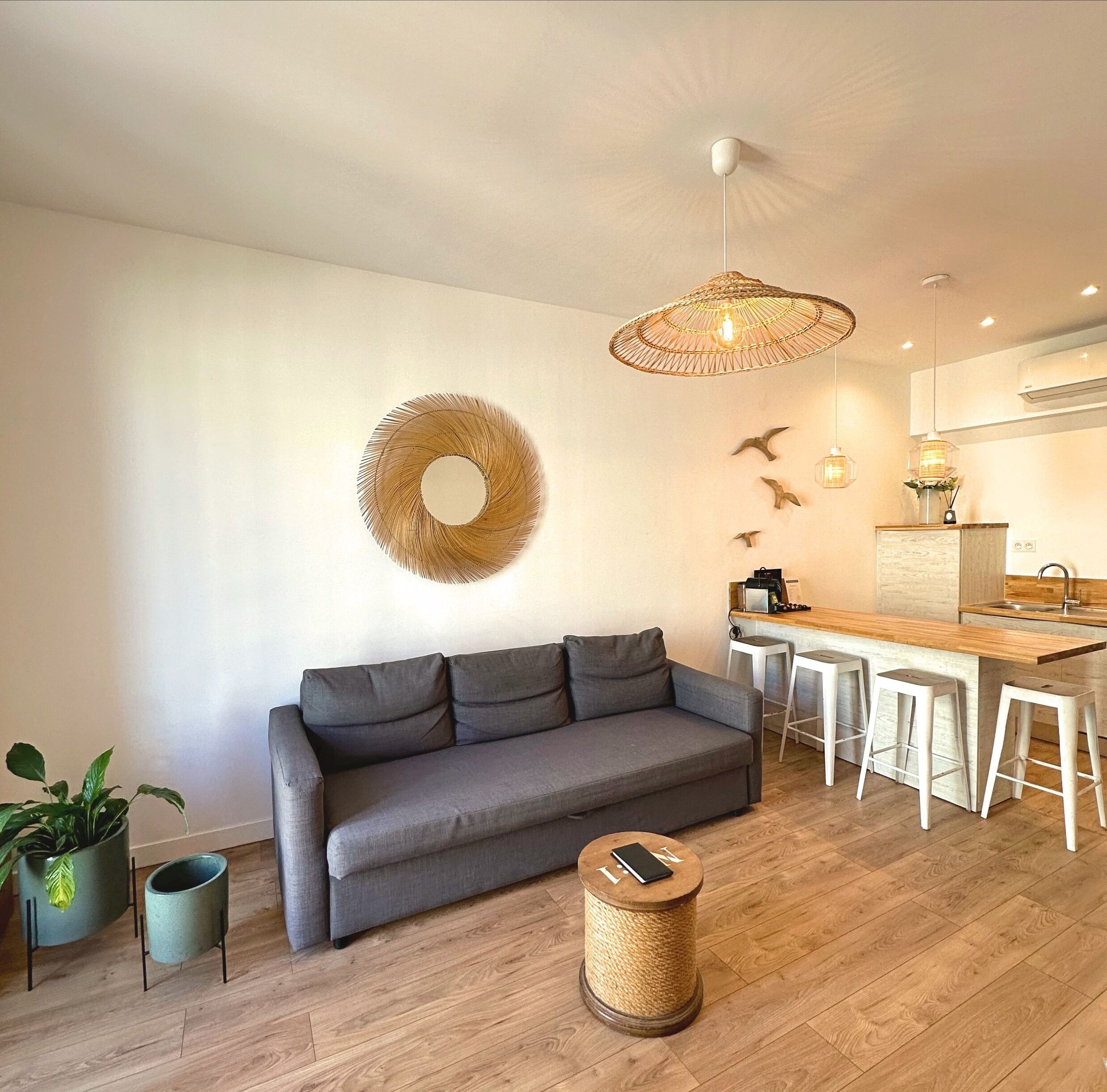 Ein modernes Wohnzimmer mit einem grauen Sofa, einem gewebten Couchtisch und einer Küchenzeile mit einer Holz Arbeitsplatte und Barhocker.