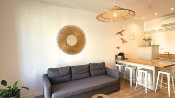A modern living room with a grey sofa, a woven coffee table, and a kitchen area with a wooden countertop and bar stools.