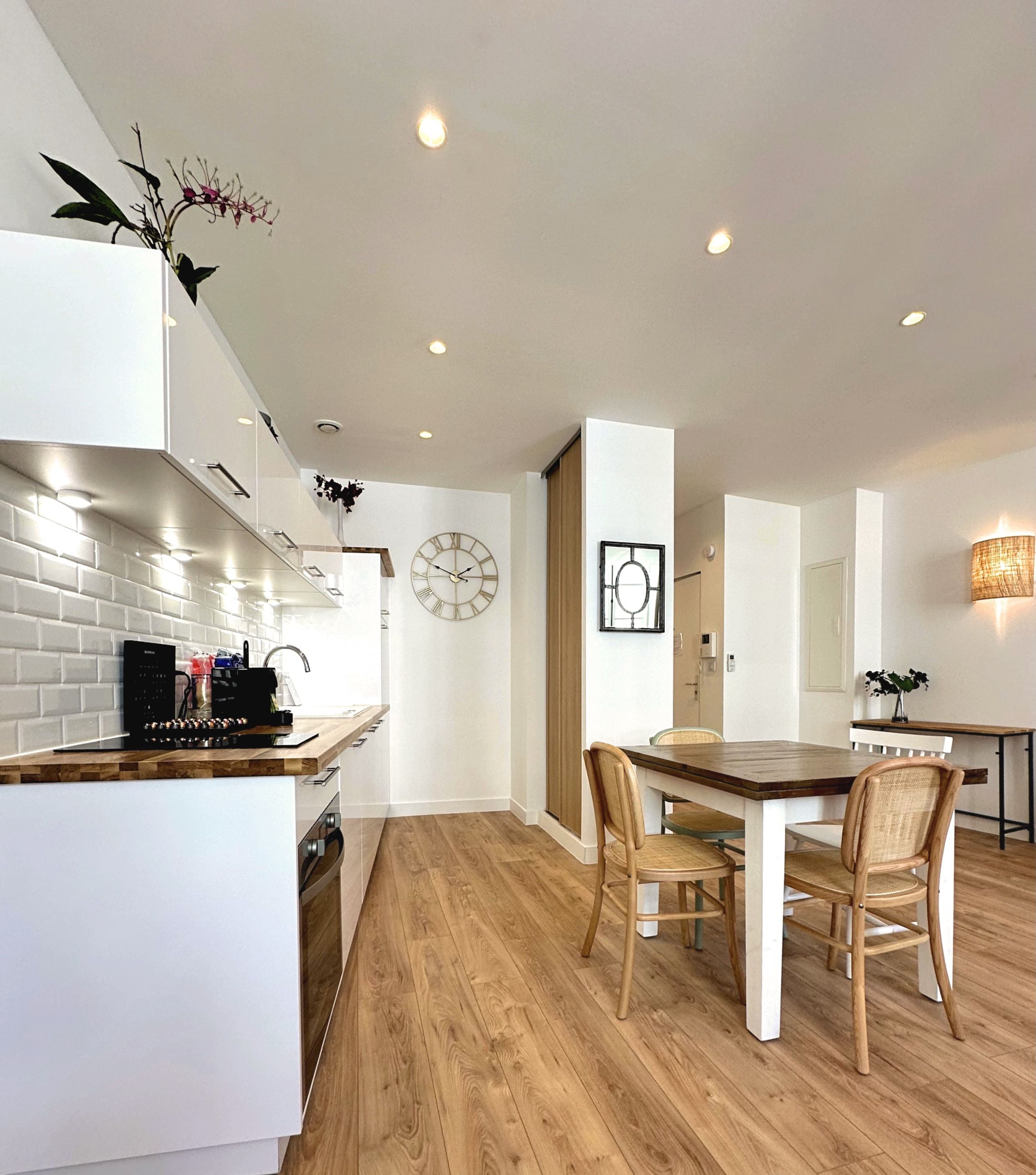 A modern kitchen with white cabinetry, a wooden dining table, and a clock on the wall.