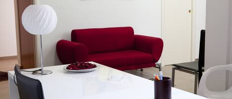 A modern living room with a white table, a red sofa, a white lamp, and a painting of lemons on the wall.