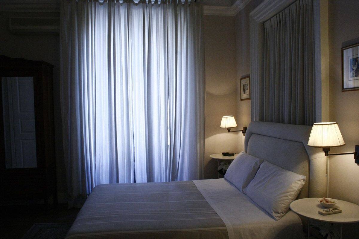 Een hotelkamer met een groot bed, twee nachtkastjes met lampen, een raam met gordijnen en een afbeelding in een lijst aan de muur.