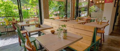 Un restaurante con mesas y sillas de madera, grandes ventanales con vistas a zonas verdes y un ventilador en el techo.