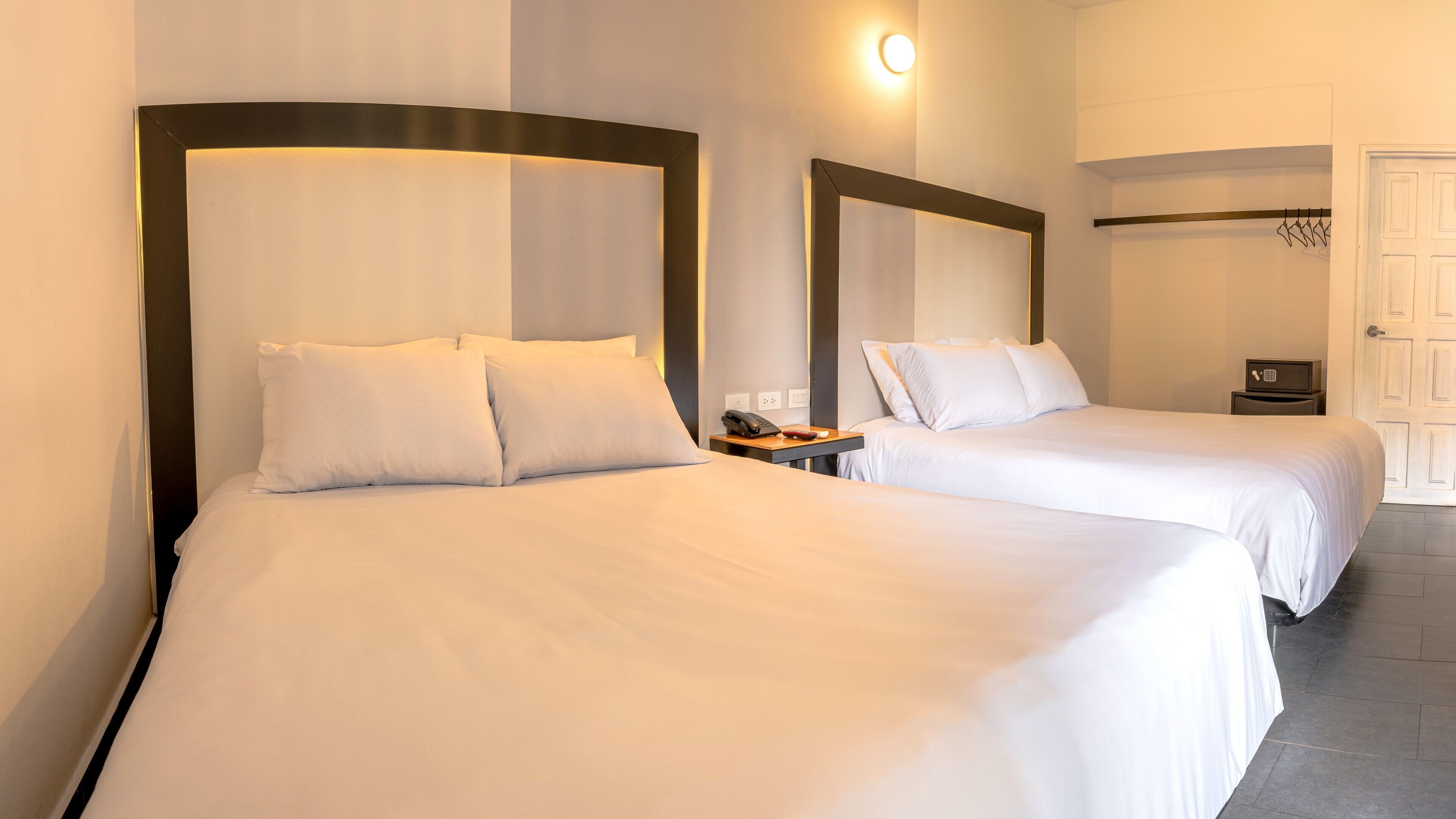 Two single beds with white linens, a bedside table with a phone and a red object, and a wall-mounted light fixture.