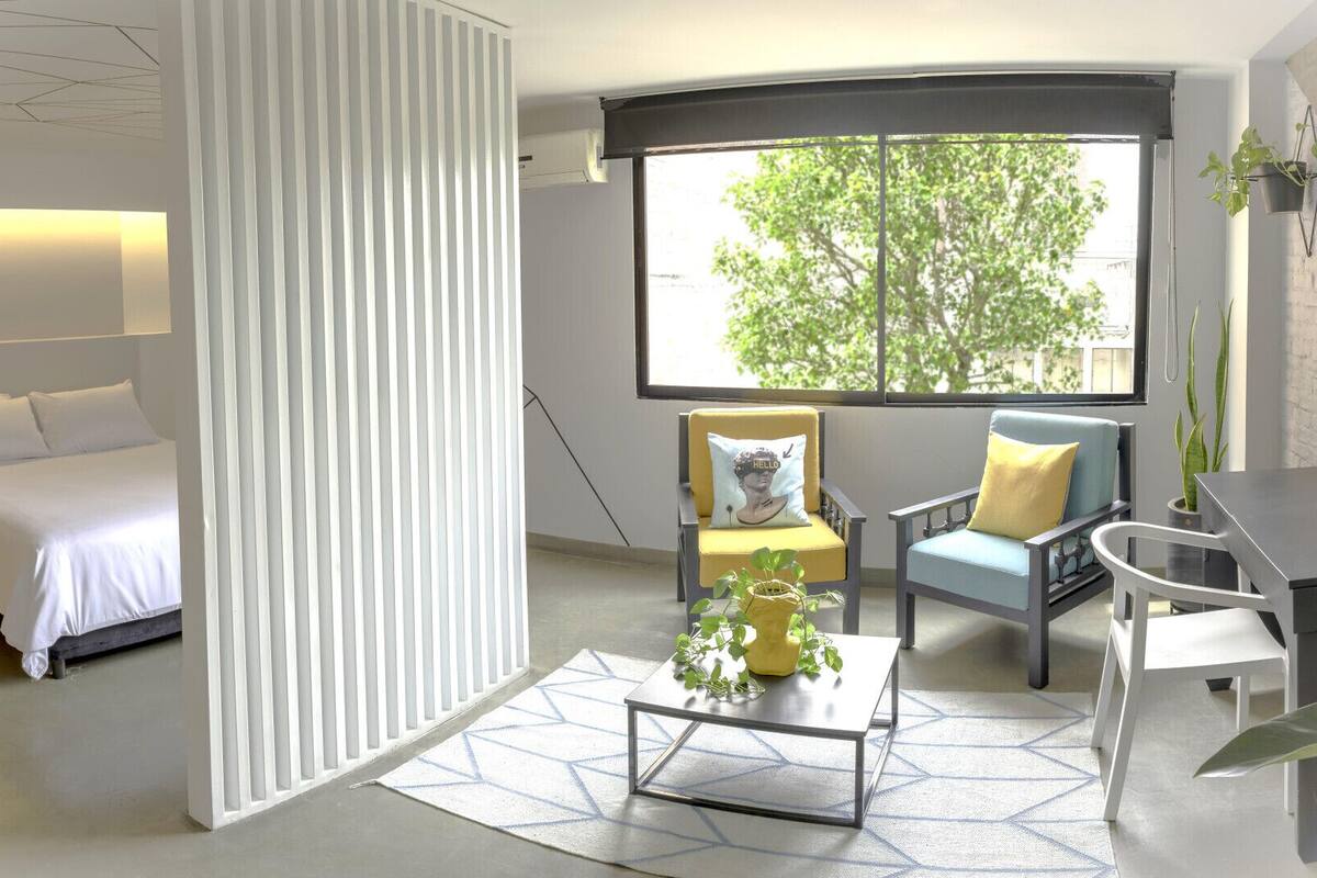 A modern hotel room with a bed, a desk, a chair, a window with a view of trees, and a white partition.