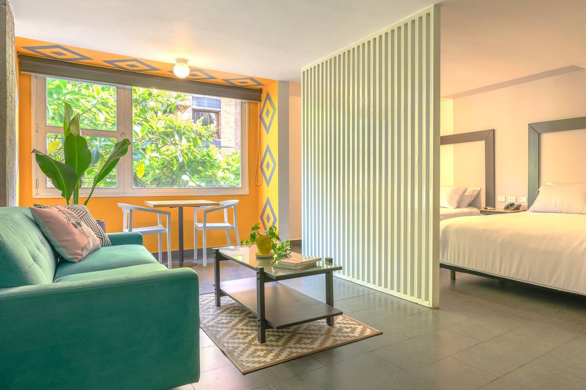 A modern hotel room with a sofa, a small table, a bed, and a large window with a view of greenery.