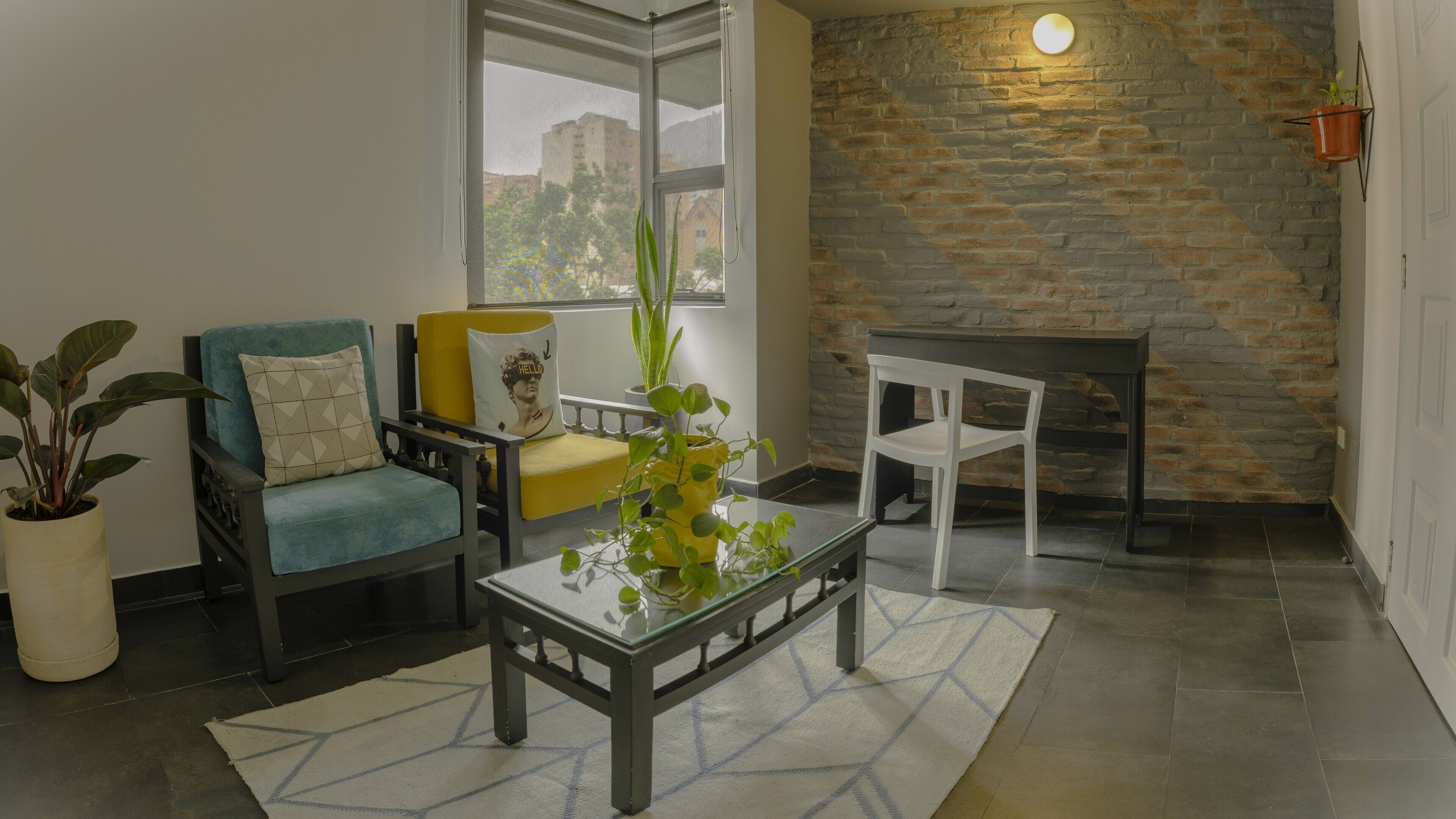 Un salon avec un canapé, un fauteuil et une petite table sur laquelle est posée une plante. Il y a un mur en briques et une fenêtre donnant sur des immeubles.