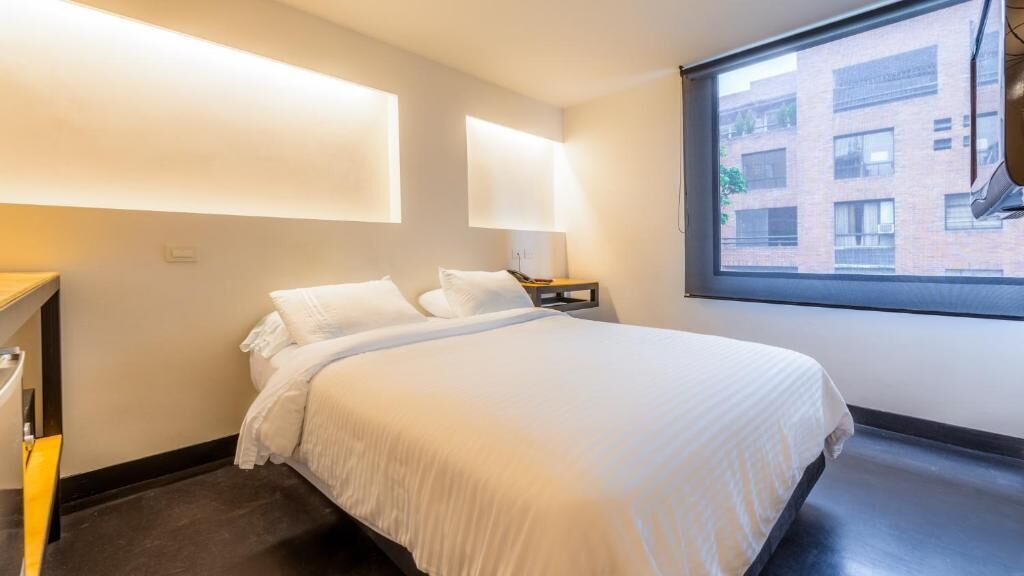 Habitación de hotel con cama, mesita de noche y ventana con vista a un edificio.
