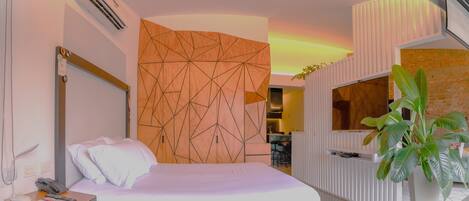 A modern hotel room with a large bed, a wooden geometric wall panel, a white console table with a phone and remote controls, and a large potted plant.