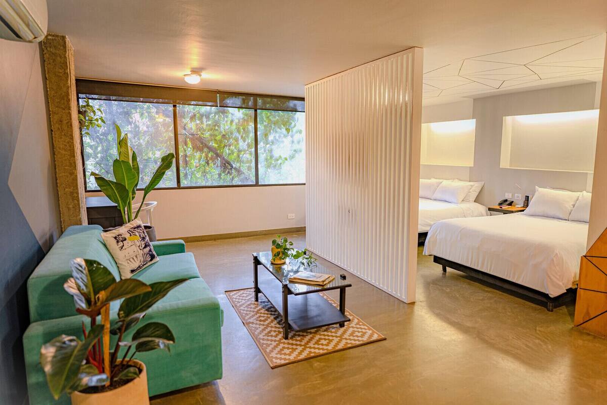 A modern hotel room with a green sofa, a bed, a desk, and a large window.