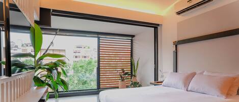 A modern hotel room with a large bed, a bedside table, a potted plant, and a window with blinds.