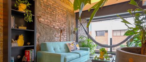 Una sala de estar con pared de ladrillo, un sofá azul, una estantería y una hamaca.