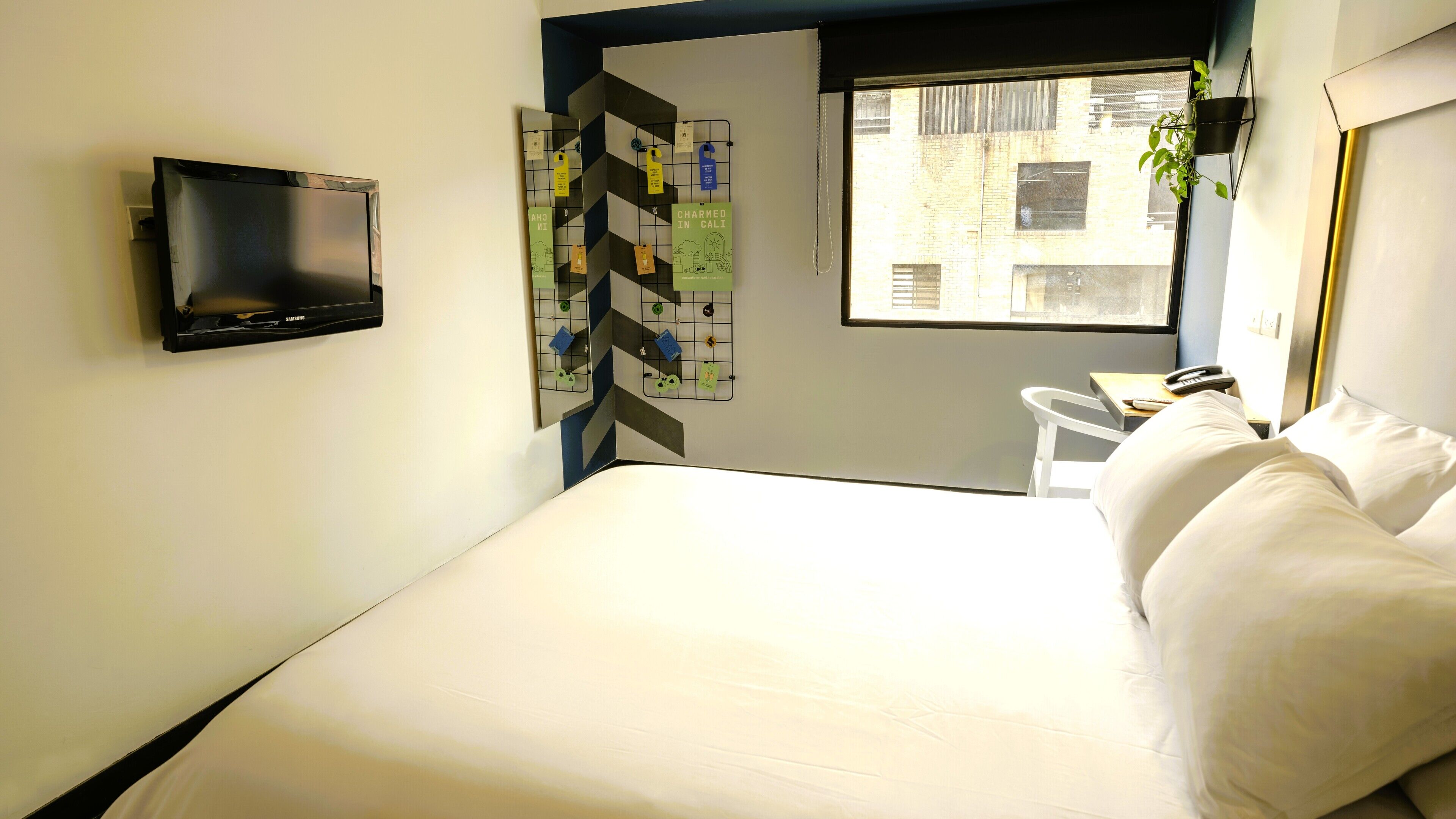 A hotel room with a bed, a flat-screen TV mounted on the wall, a small table with a chair, and a window with a view of buildings.