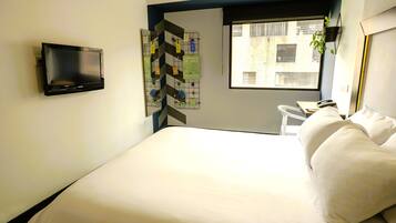 A hotel room with a bed, a flat-screen TV mounted on the wall, a small table with a chair, and a window with a view of buildings.
