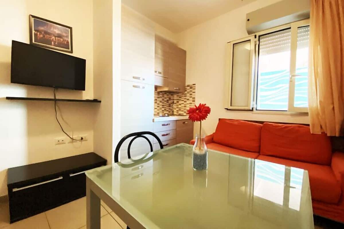Un espace de vie compact comprenant une table à manger en verre, un canapé, un coin cuisine et un téléviseur fixé au mur.