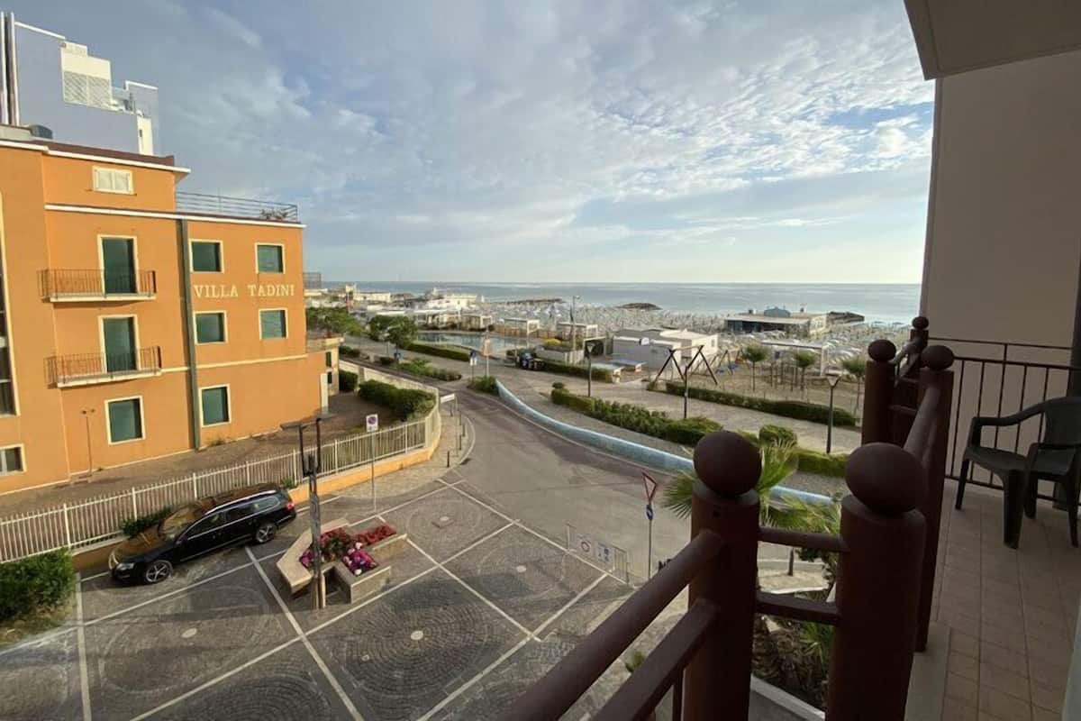 Vue de la villa Tadini, avec un espace de stationnement, une route menant à la mer et un ciel dégagé.