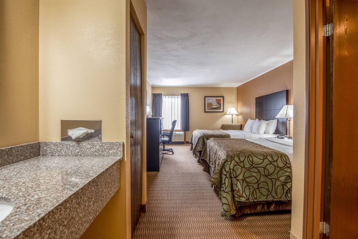 A hotel room with a granite countertop, two beds with green patterned bedspreads, a desk with a chair, and a painting on the wall.
