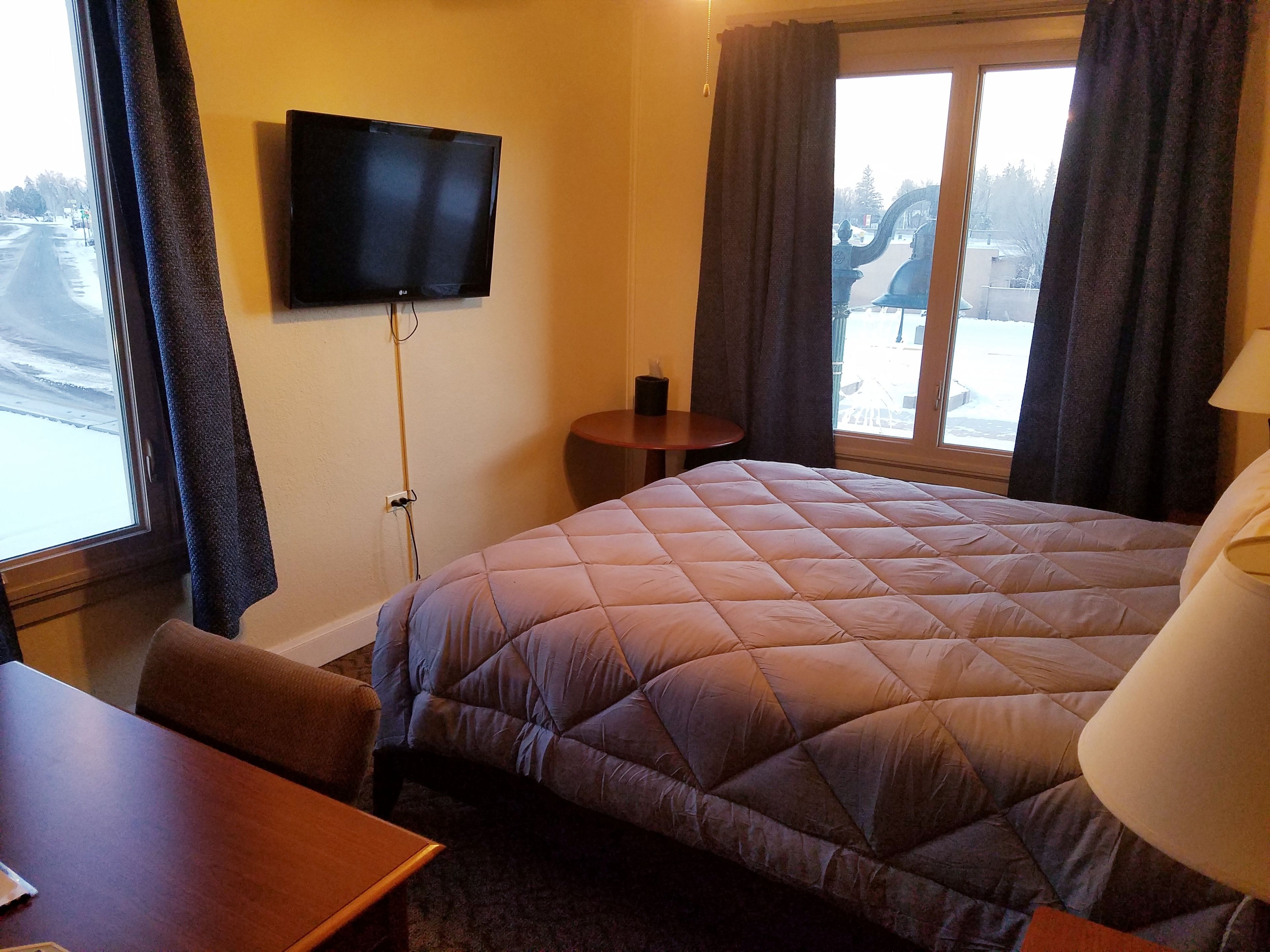 Habitación de hotel con una cama grande, un televisor montado en la pared, una mesita y una ventana con cortinas.