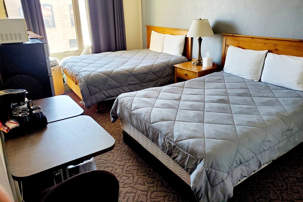 A hotel room with two beds, a desk, and a chair. There is a lamp on the desk and a window with curtains.