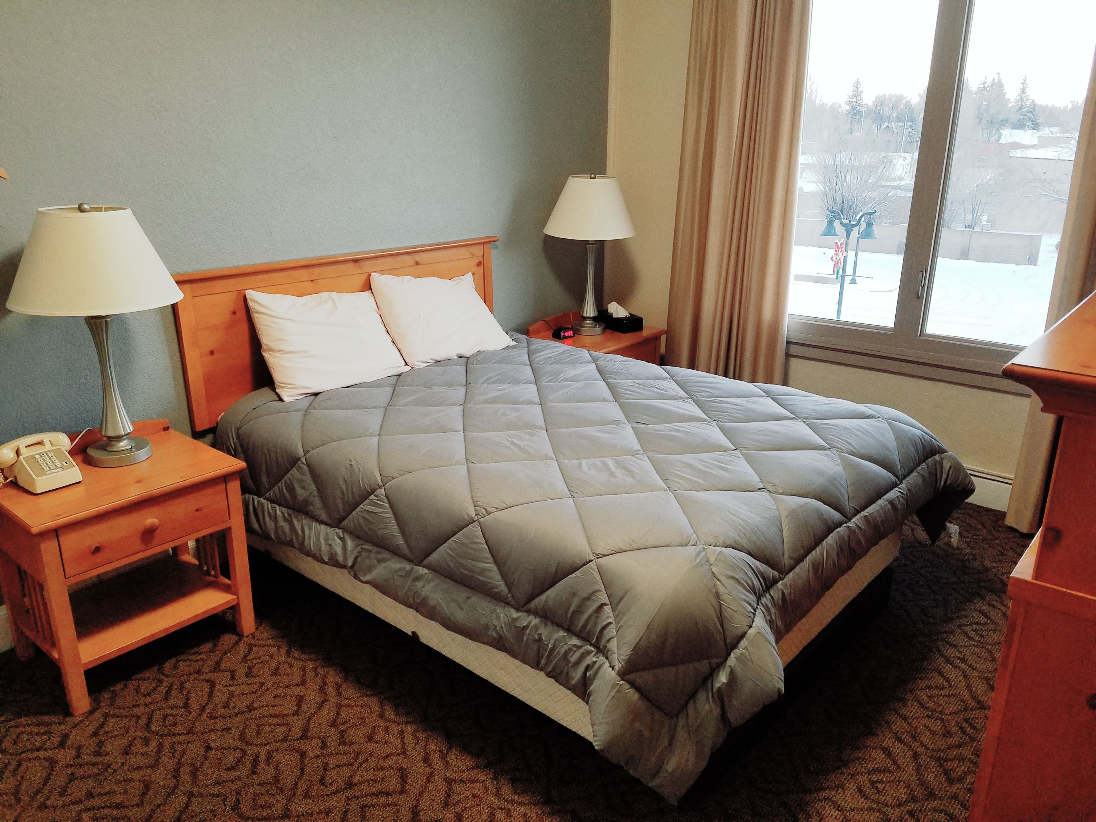 A neatly made bed with a grey quilt, two bedside lamps, a nightstand with a telephone, and a window with curtains.