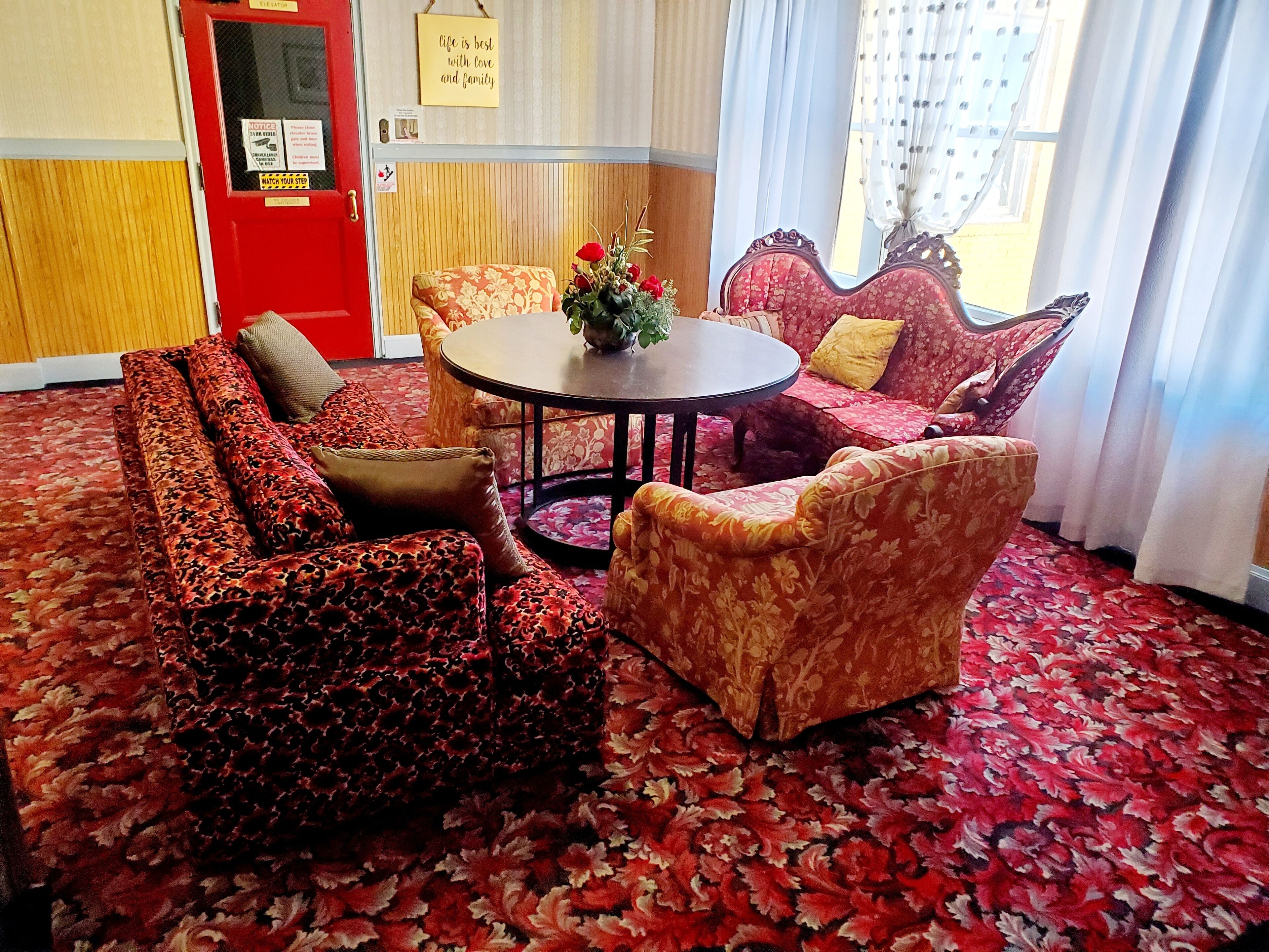 Une pièce avec une porte rouge, des canapés à motifs floraux, une table ronde et une fenêtre ornée de rideaux légers.