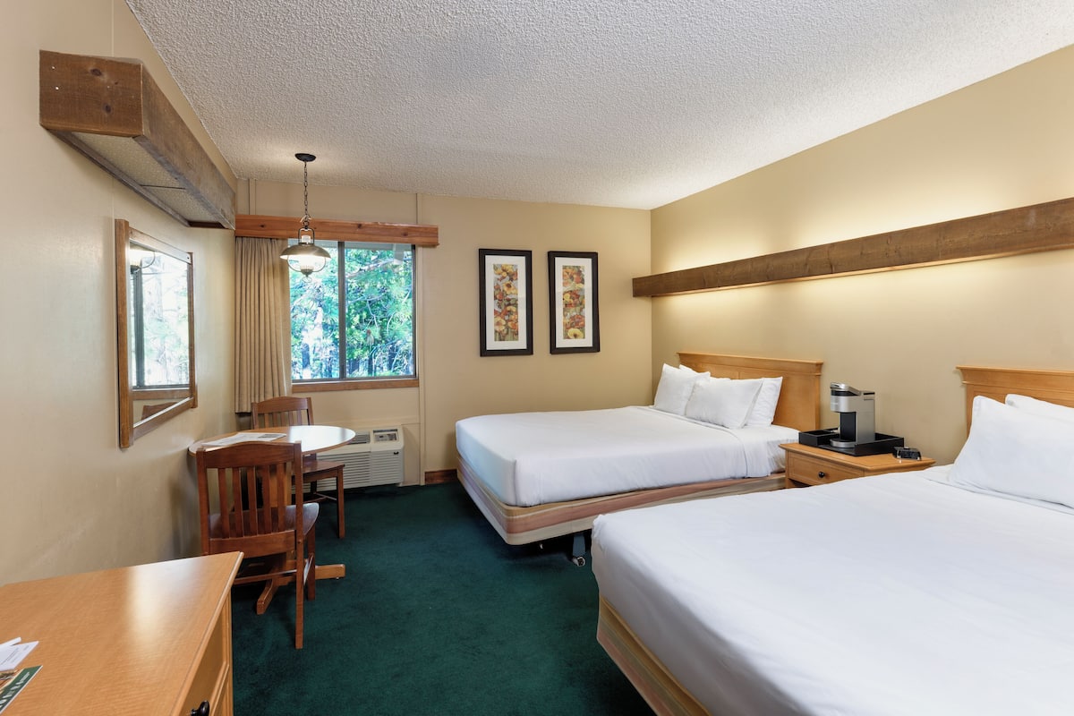 Une chambre d’hôtel avec deux lits, un bureau, une chaise, une fenêtre donnant sur des arbres et un ventilateur de plafond.