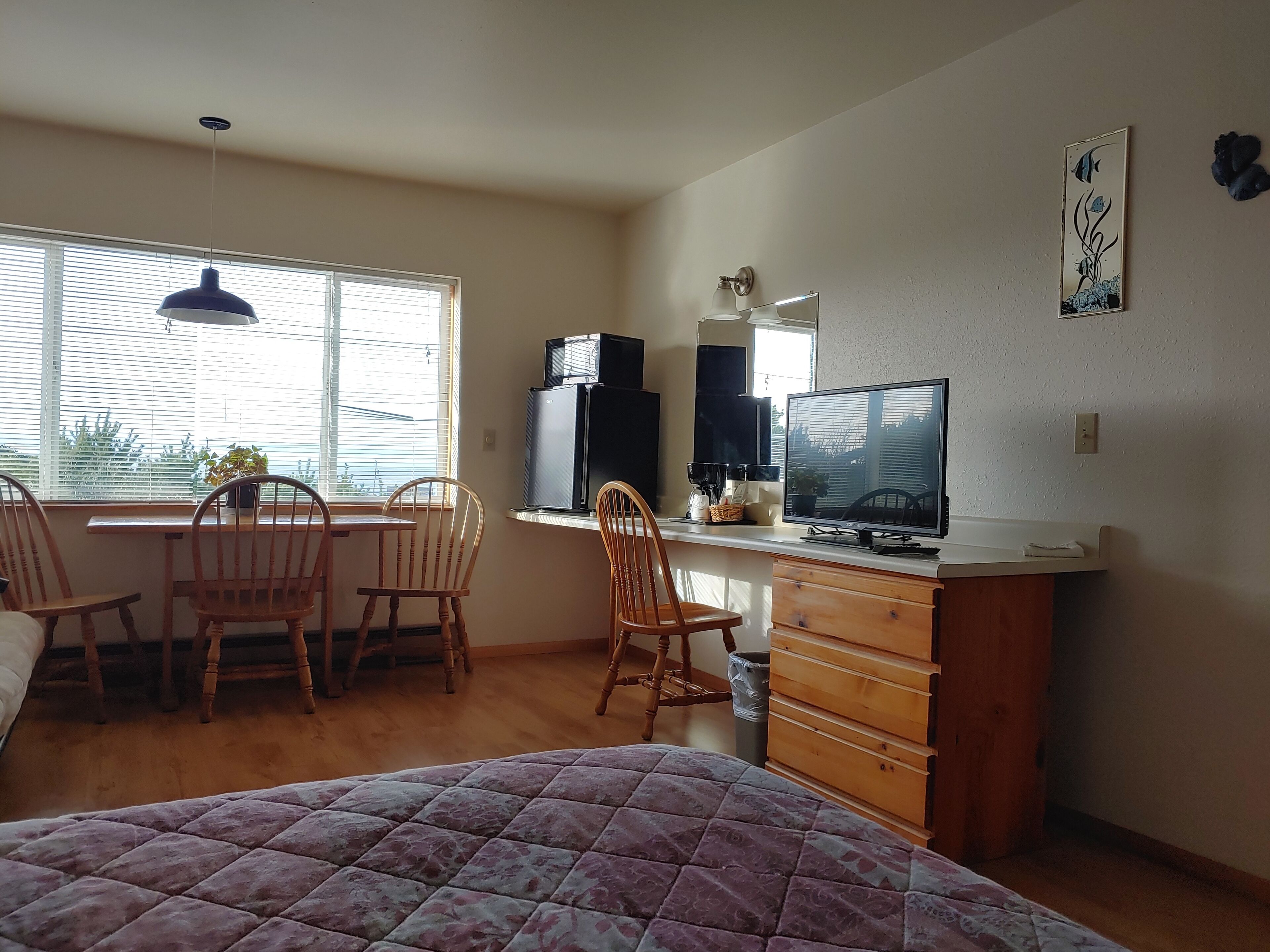 A compact room with a bed, a desk with a computer, two chairs, a small cabinet, and a window with blinds.