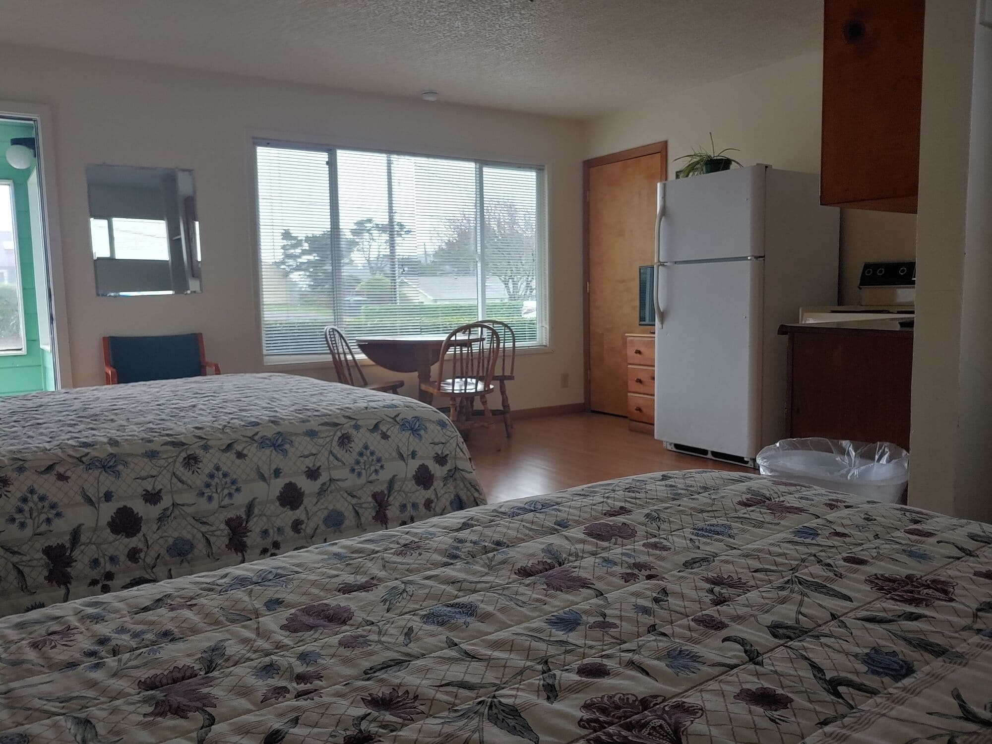 A room with two beds, a dining table with chairs, a refrigerator, and a window with blinds.