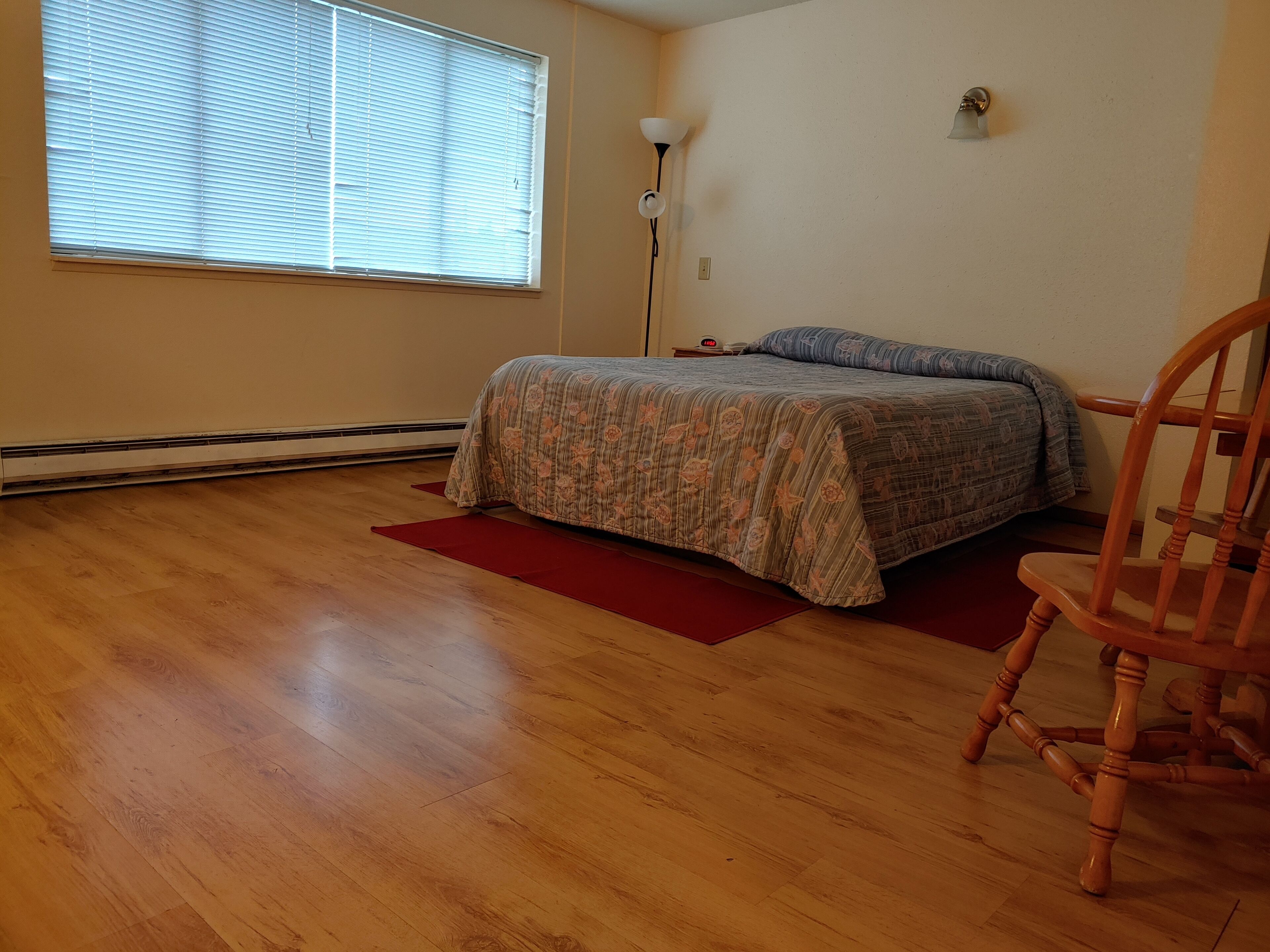 Un dormitorio con piso de madera, una cama con ropa de cama floral, un tapete rojo, una silla de madera, una lámpara de pie y una ventana con persianas.