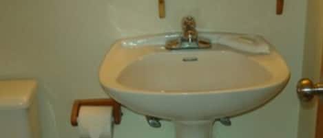 A bathroom with a white sink, toilet, and trash can. There is a mirror above the sink and a small shelf with a tissue box.
