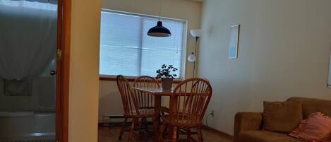 A cozy living room with a brown sofa, a dining table with chairs, a large window with blinds, and a ceiling light.