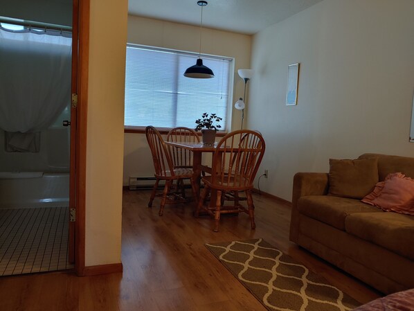 A cozy living room with a brown sofa, a dining table with chairs, a large window with blinds, and a ceiling light. - The Dublin House Motel (Yachats)