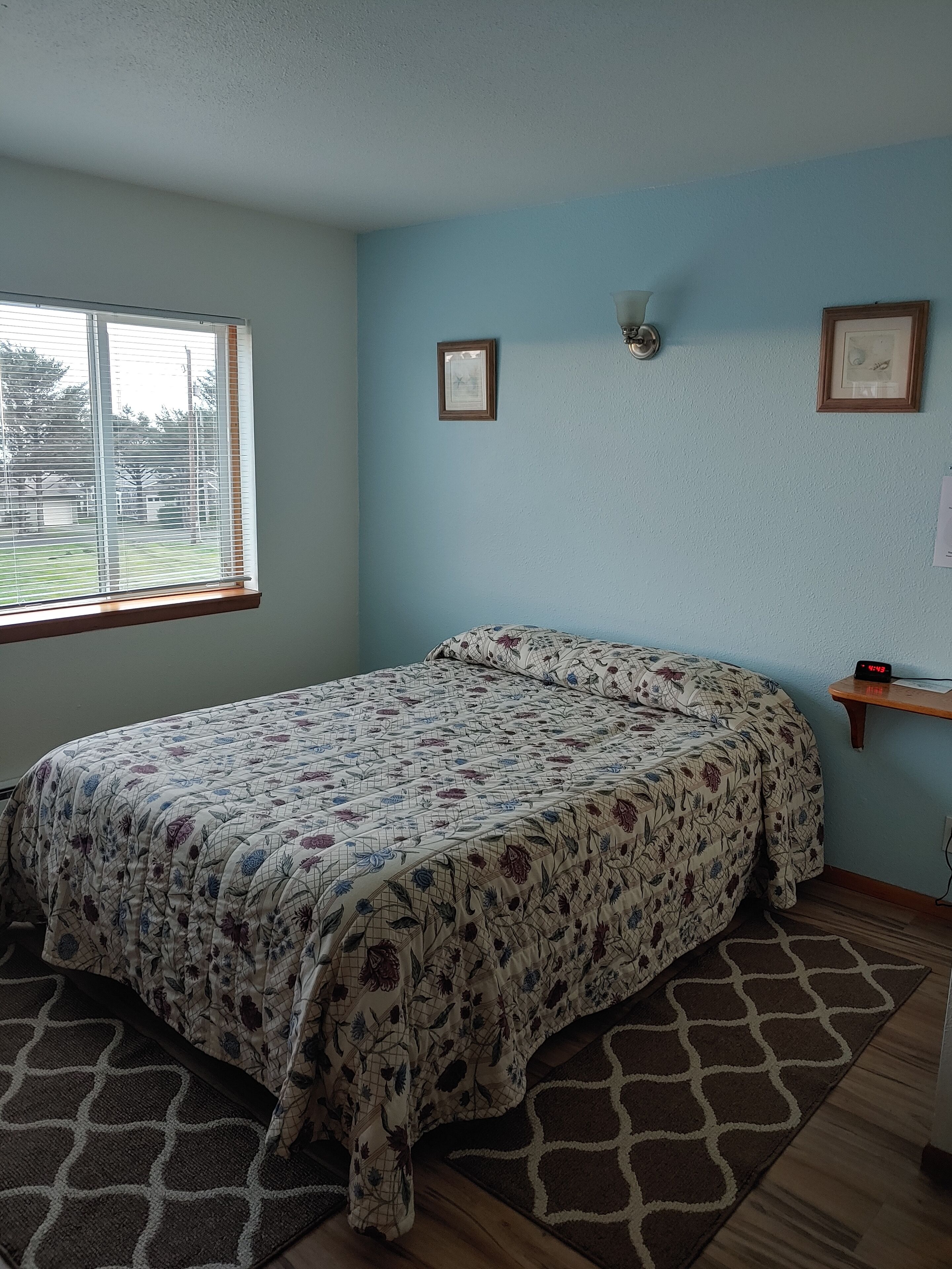 Un dormitorio con colcha estampada, una ventana con persianas, un reloj de pared y cuadros enmarcados.