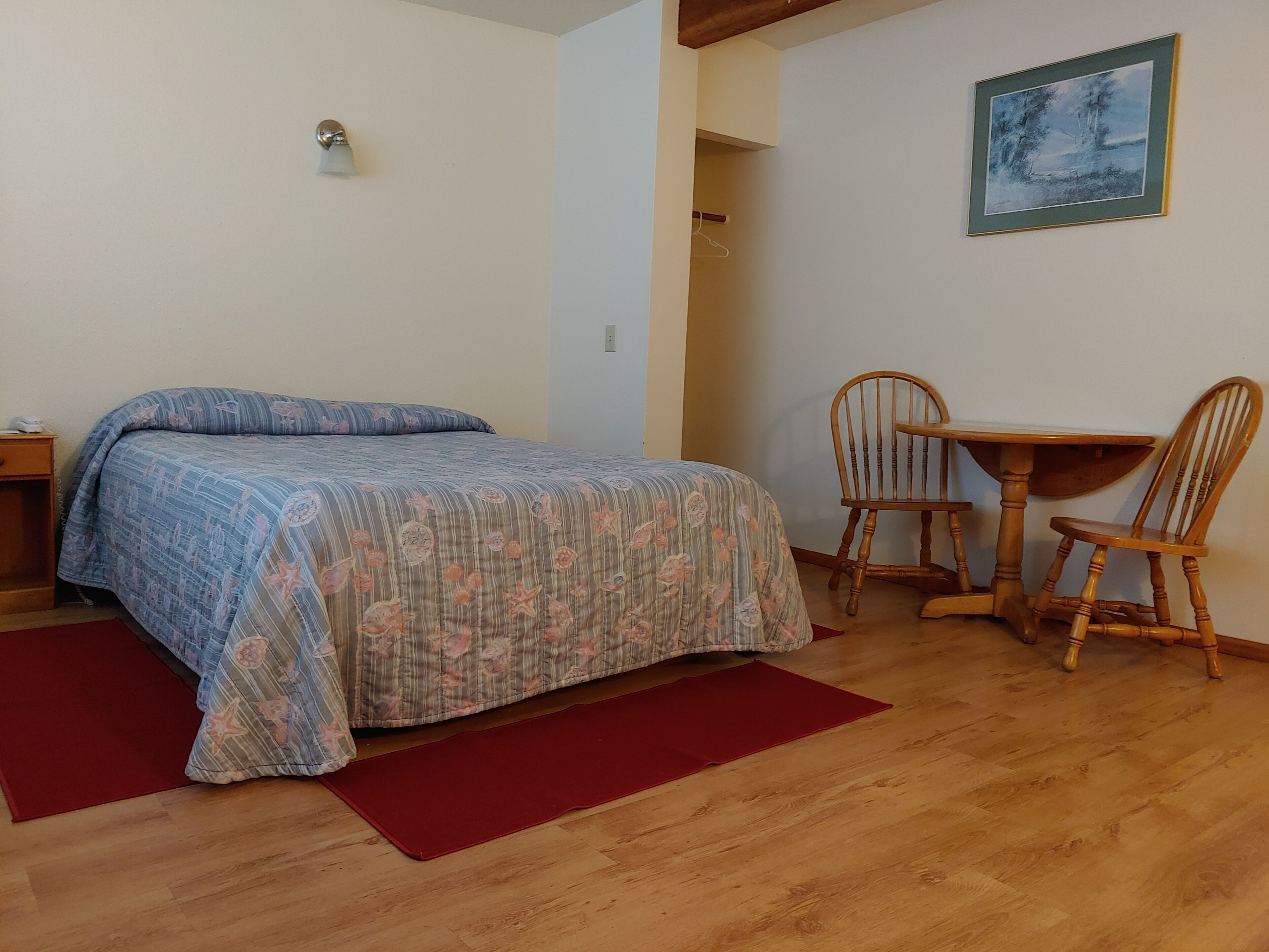 Quarto com cama, mesa de madeira com duas cadeiras, tapete vermelho e um quadro na parede.