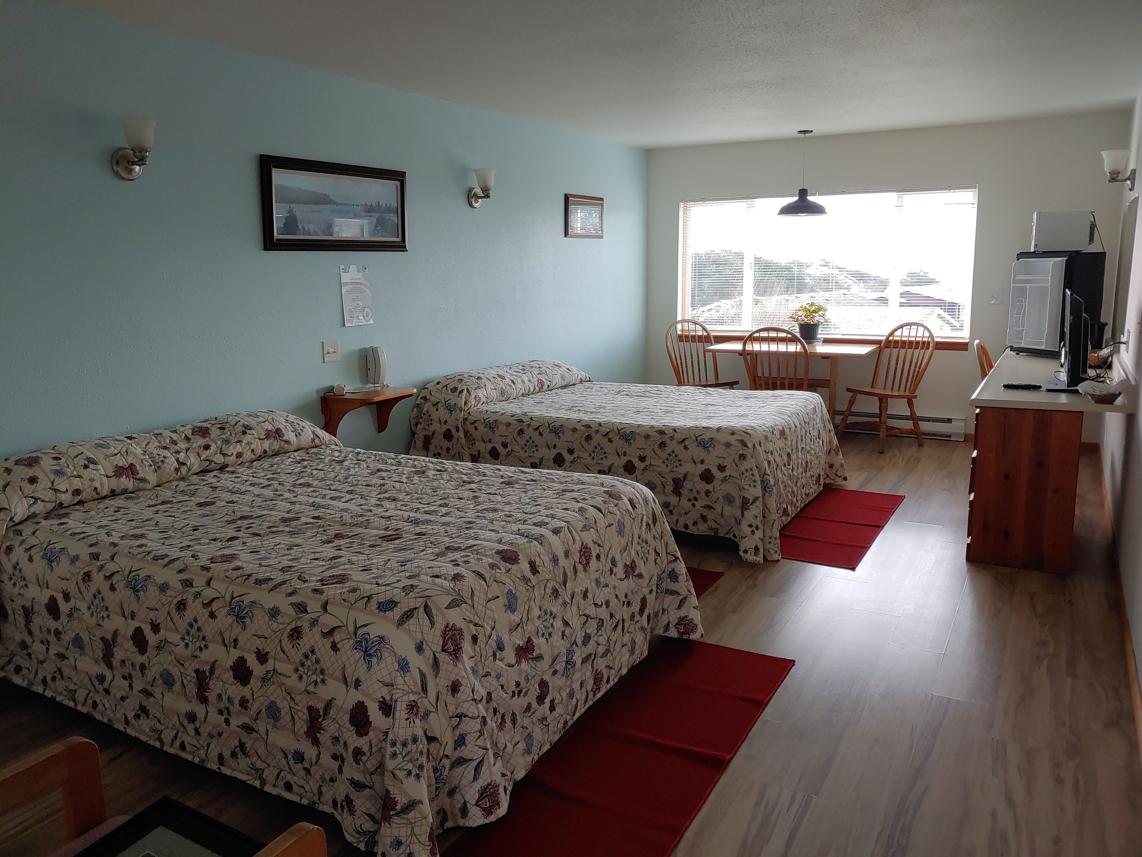 A room with two beds, a desk, a chair, a TV, and a window with blinds.