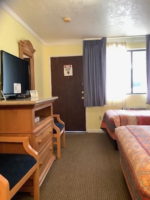 A hotel room with a bed, a desk with a computer, a TV, a door with a sign, and a window with curtains.