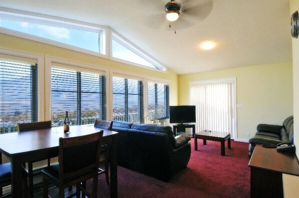 A living room with a dining area, large windows, a flat-screen TV, and a ceiling fan.