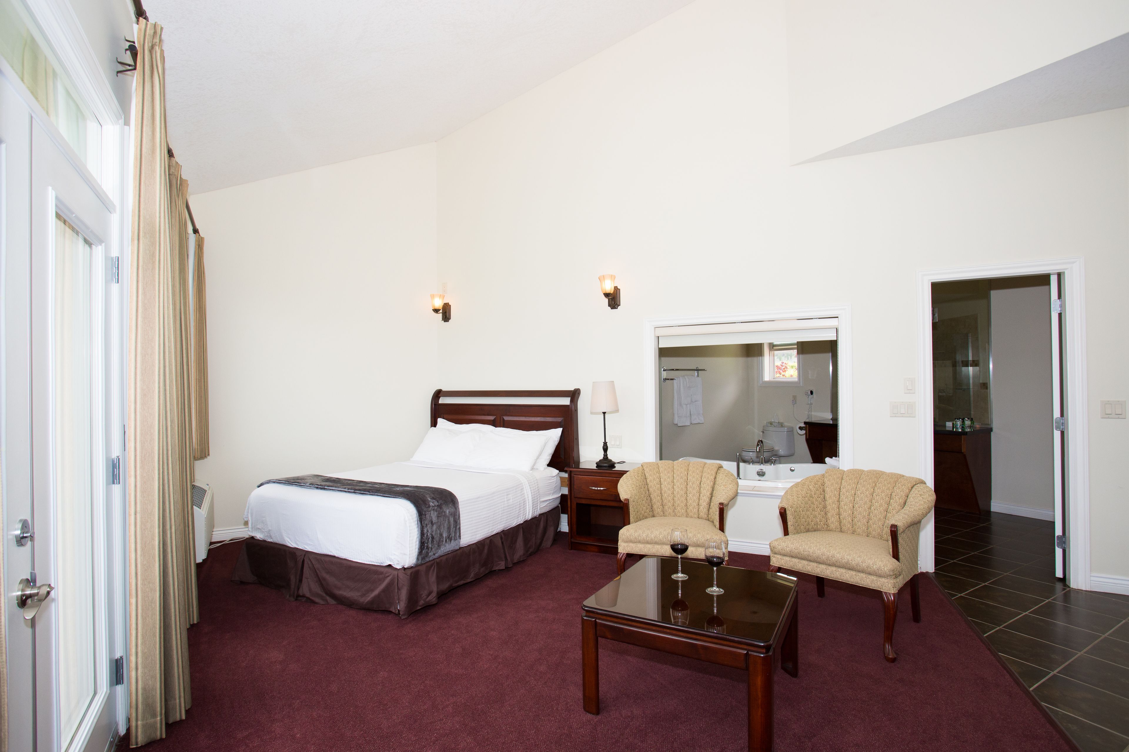 Habitación de hotel con una cama, dos sillones, una mesita de centro, una mesita de noche, una lámpara y un baño visible a través de una puerta abierta.