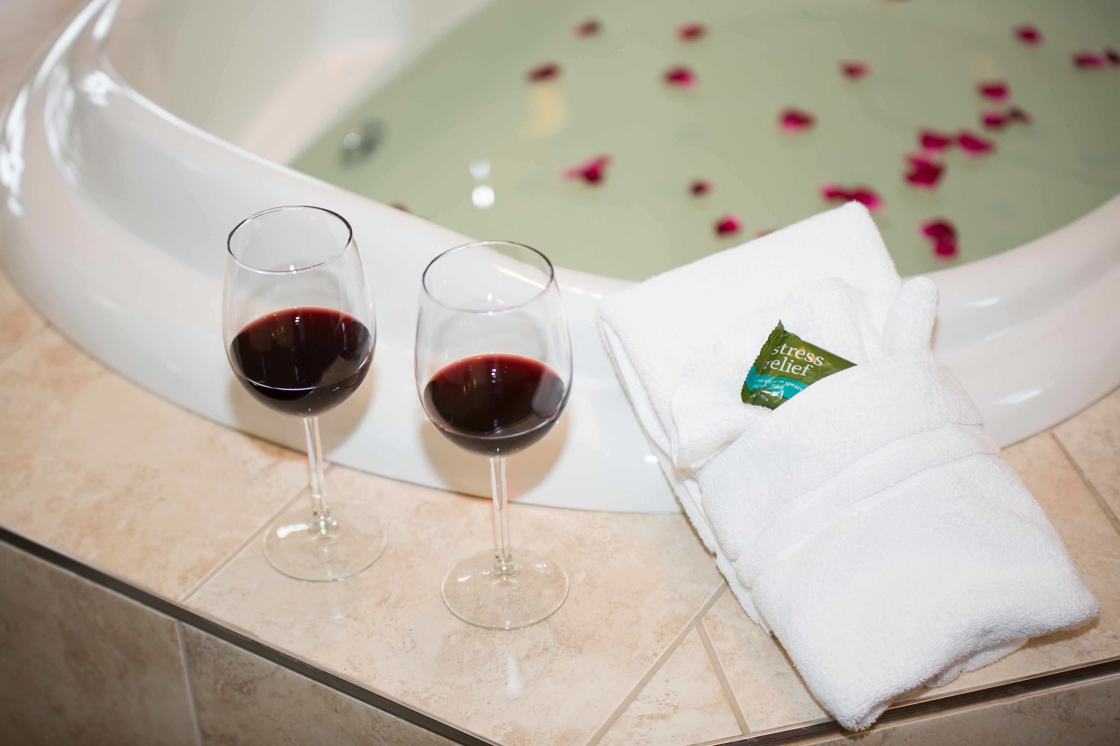 Una bañera con pétalos de rosa, dos copas de vino tinto y una toalla blanca con un lazo verde.